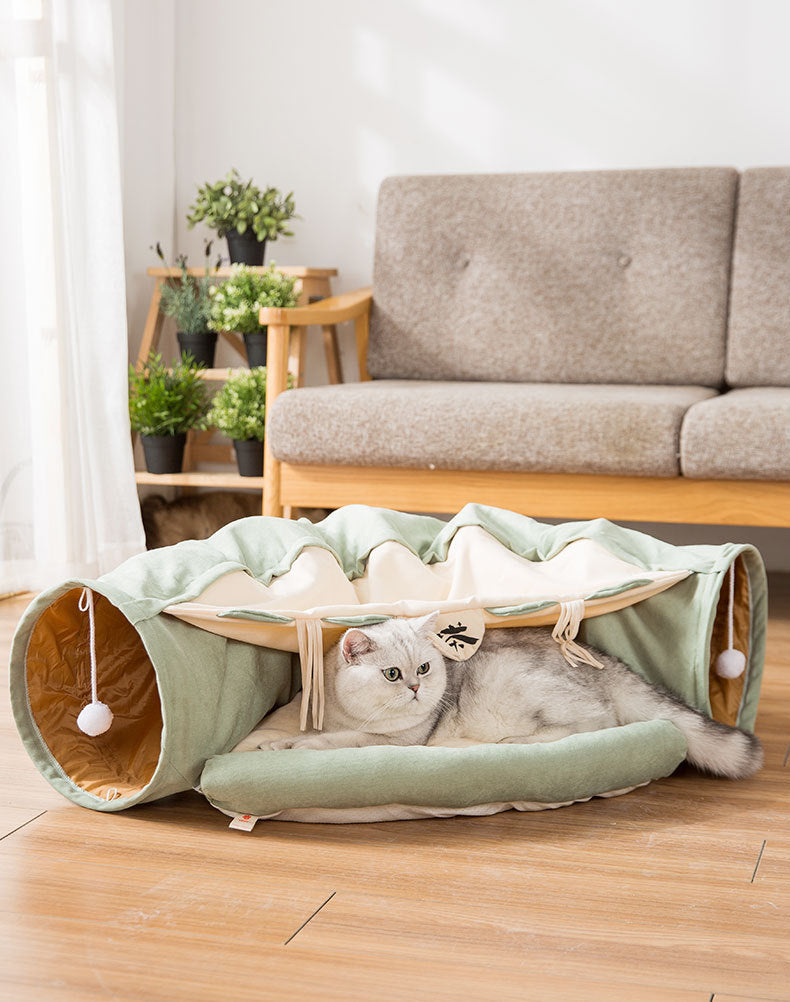 Gray cat resting inside a collapsible green cat tunnel with hanging toys on a wooden floor in a living room.