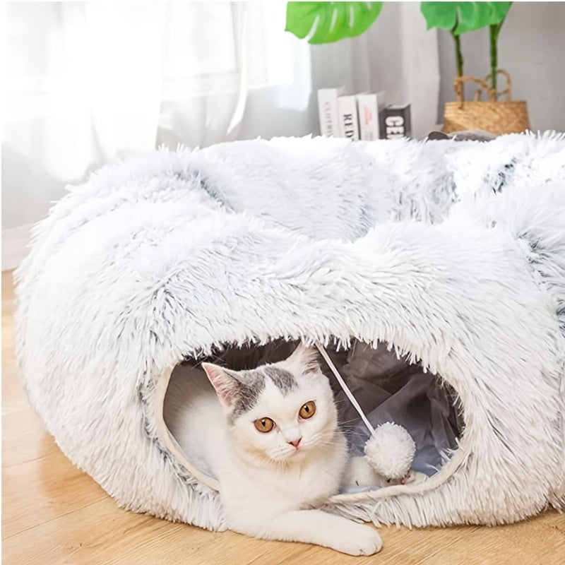 White fluffy foldable cat tunnel nest with a small playful cat inside on wooden floor near plants and books.